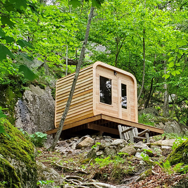 A Dundalk LeisureCraft luxury cabin, the Dundalk Canadian Timber Luna 2 to 3 Person White Cedar, sits on top of a rock in the woods, made with Eastern White Cedar.